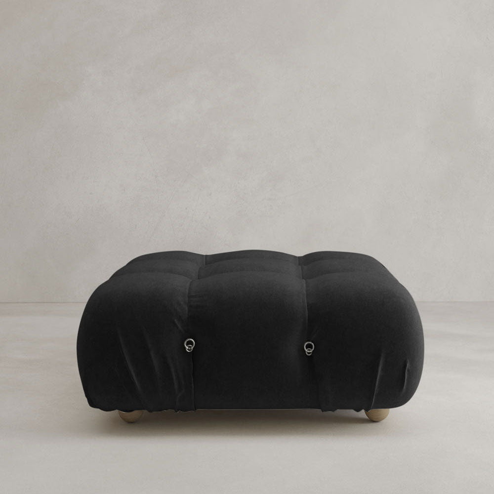 The Belmont Ottoman, featuring a black tufted design, rounded edges, and small wooden feet, sits on a light neutral floor against a plain beige wall—an ideal match for iconic classic or modular sofas.