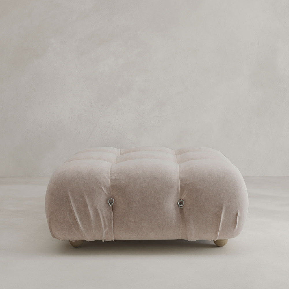 The Belmont Ottoman, large and beige with a tufted, velvety texture and classic style, sits on a light-colored floor against a plain light grey wall.