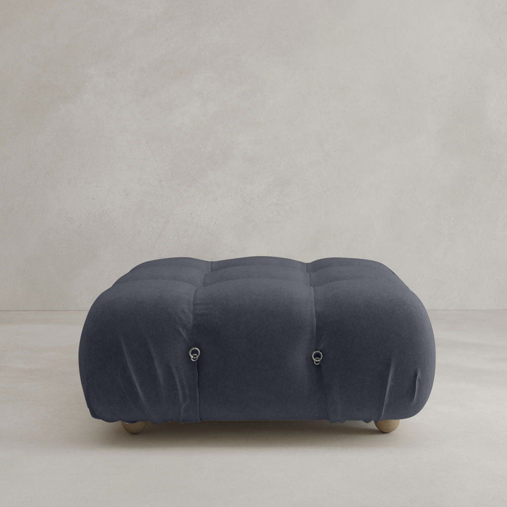 The Belmont Ottoman, featuring dark blue velvet, tufted buttons, and rounded edges, brings iconic classic charm to a light beige floor set against a plain, light-colored wall.