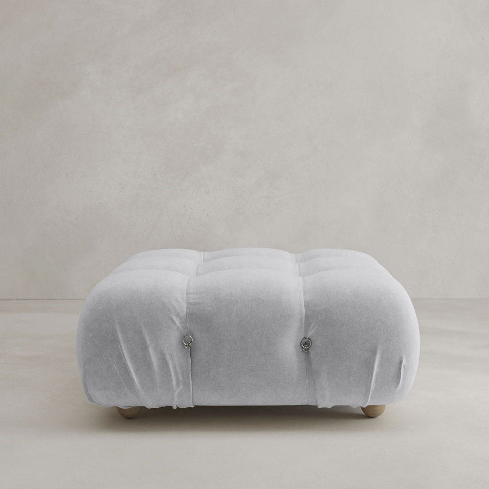 The Belmont Ottoman is a soft, square ottoman in light gray with button-tufted details and rounded edges, featuring a modular design that rests seamlessly on a neutral background.