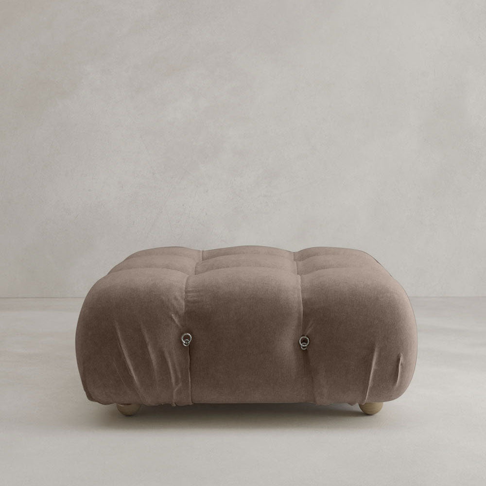 The Belmont Ottoman is a large, square, brown tufted piece with soft upholstery and round wooden feet, featuring a modular design. It stands out beautifully against a light grey textured wall and floor.