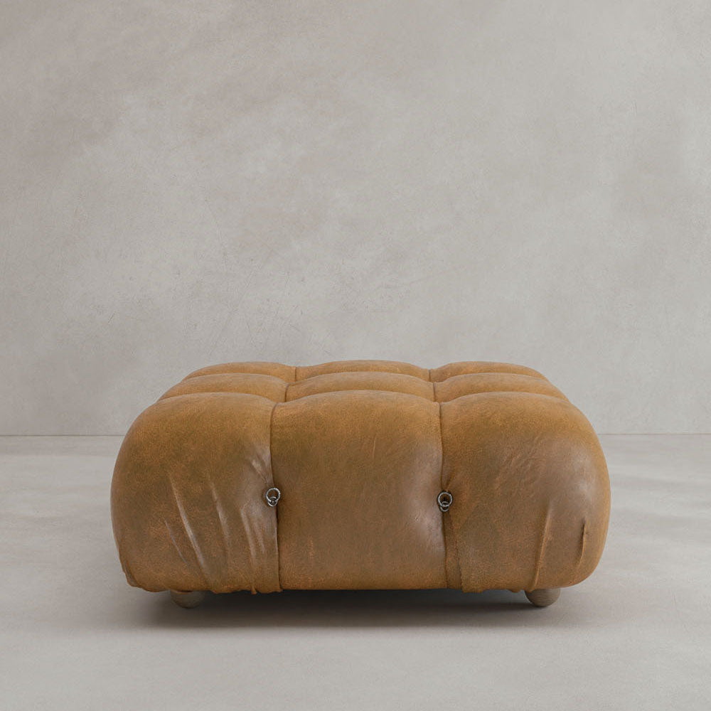 The Belmont Ottoman, a square tufted leather piece in light brown with a modular design, sits on a light gray floor against a plain, light-colored wall.