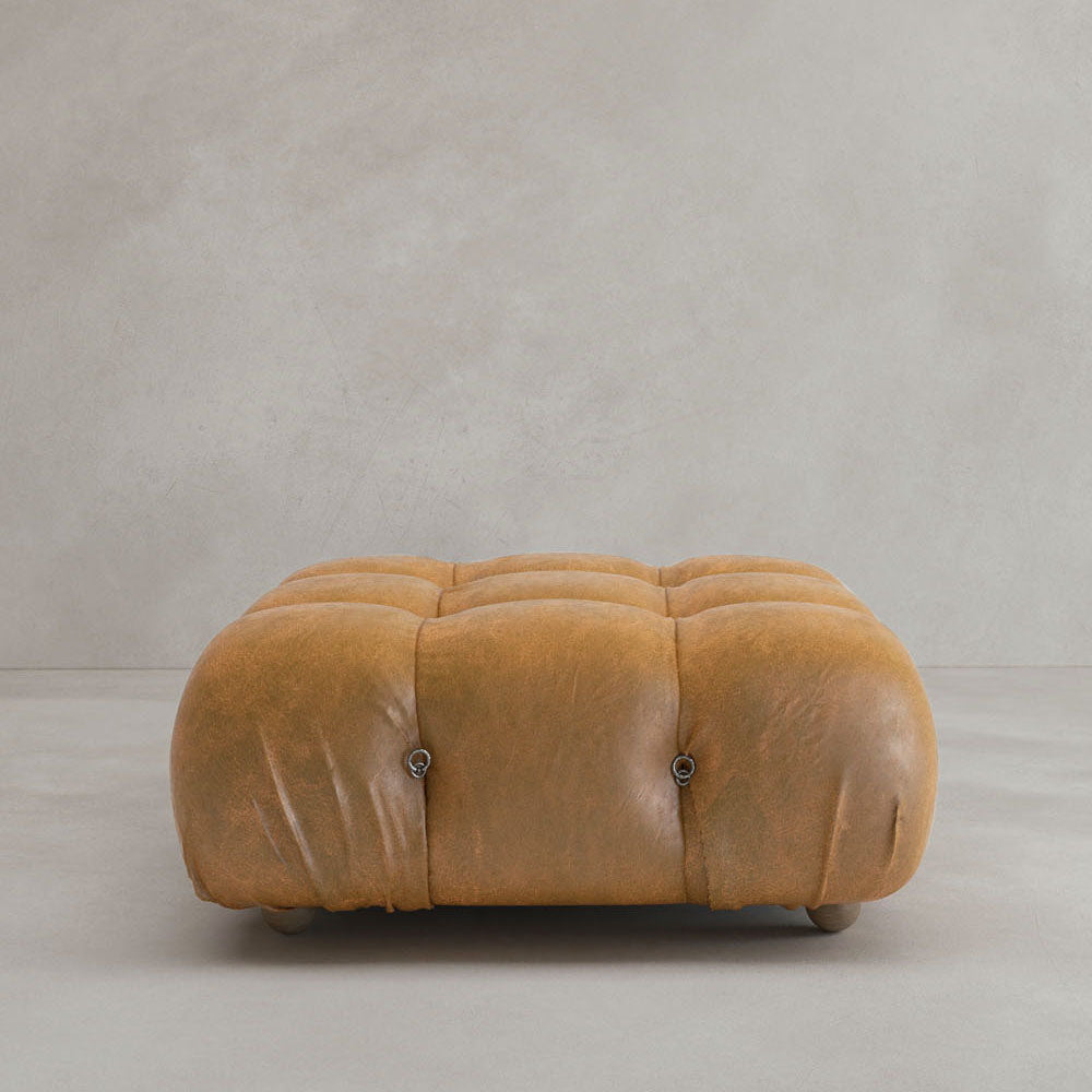 The Belmont Ottoman, a rectangular tan leather piece with a tufted top and rounded corners, features modular design. It sits on a light gray floor against a plain gray wall.