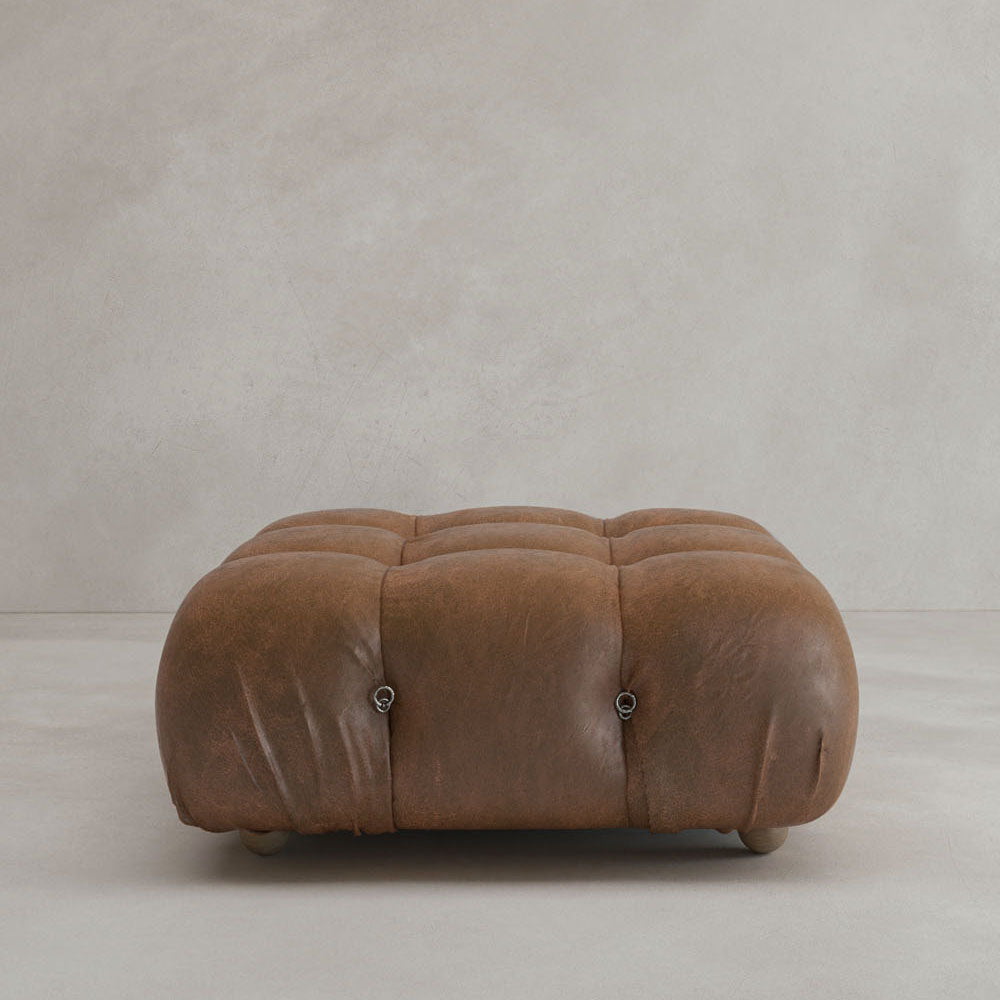 The Belmont Ottoman is a square, brown, tufted leather piece with subtle creasing and small wooden feet, set against a plain light grey background. It showcases modular design inspired by iconic classic furniture.