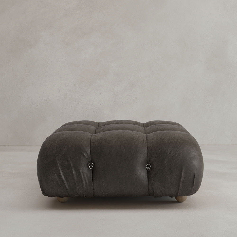 The Belmont Ottoman is a low, square, tufted dark gray piece with rounded edges and subtle button accents, highlighting modular design on a light minimalist background.
