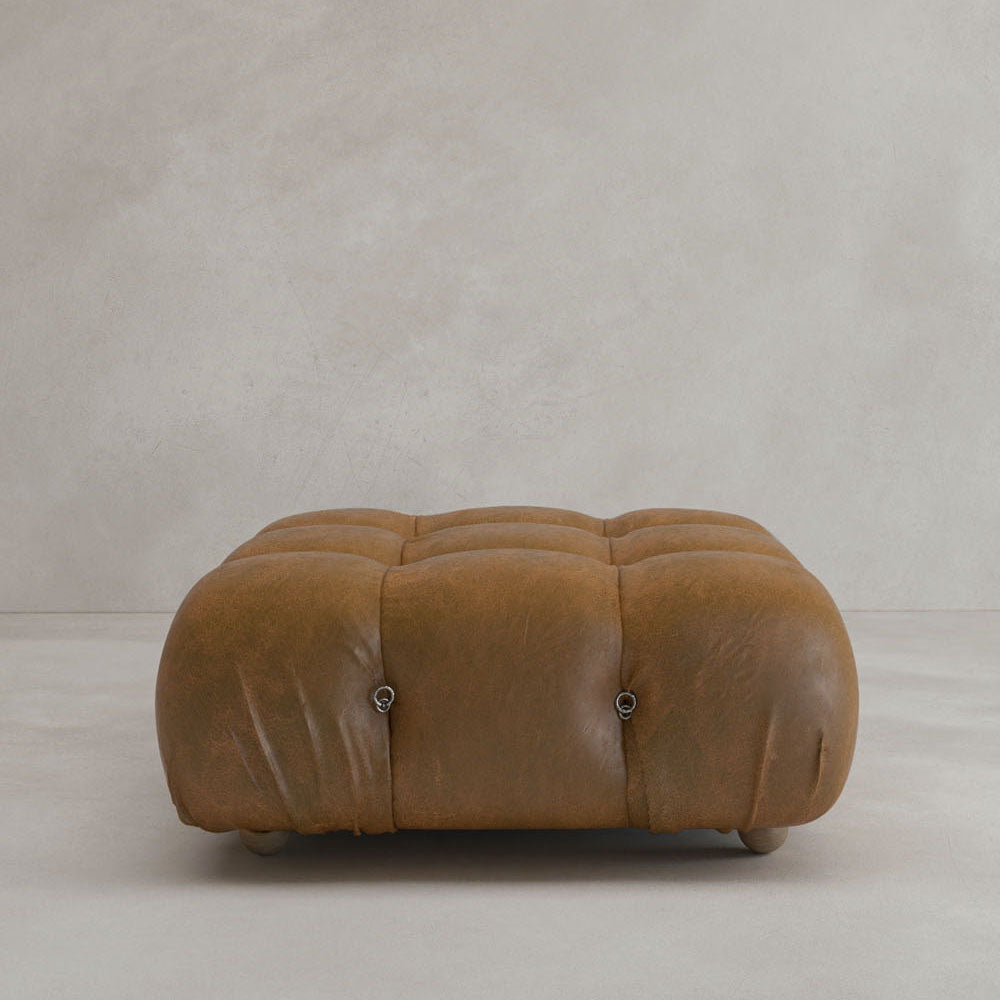 The Belmont Ottoman is a low, rectangular, tufted brown leather piece with rounded edges and button details, highlighting modular design as it sits on a light-colored floor against a plain light gray wall.