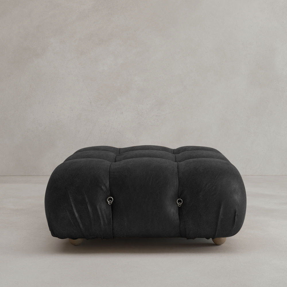 The Belmont Ottoman, a black, tufted piece with soft plush texture, sits on a light floor by a gray wall. Its modular design with rounded edges and small wooden feet make this iconic classic both stylish and charming.