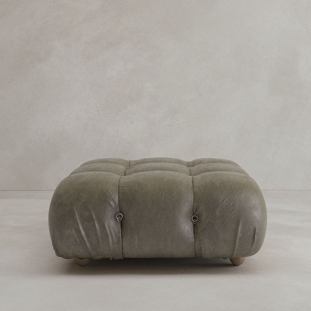 The Belmont Ottoman, a square tufted olive-green leather piece with round wooden feet, sits on a light floor against a neutral wall and pairs perfectly with a cloud-like sofa for an iconic classic look.