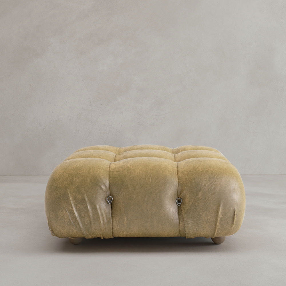 The Belmont Ottoman, a large square piece in tan leather with tufted buttons and rounded edges, brings classic style to the space as it sits on a light gray floor against a plain gray wall.