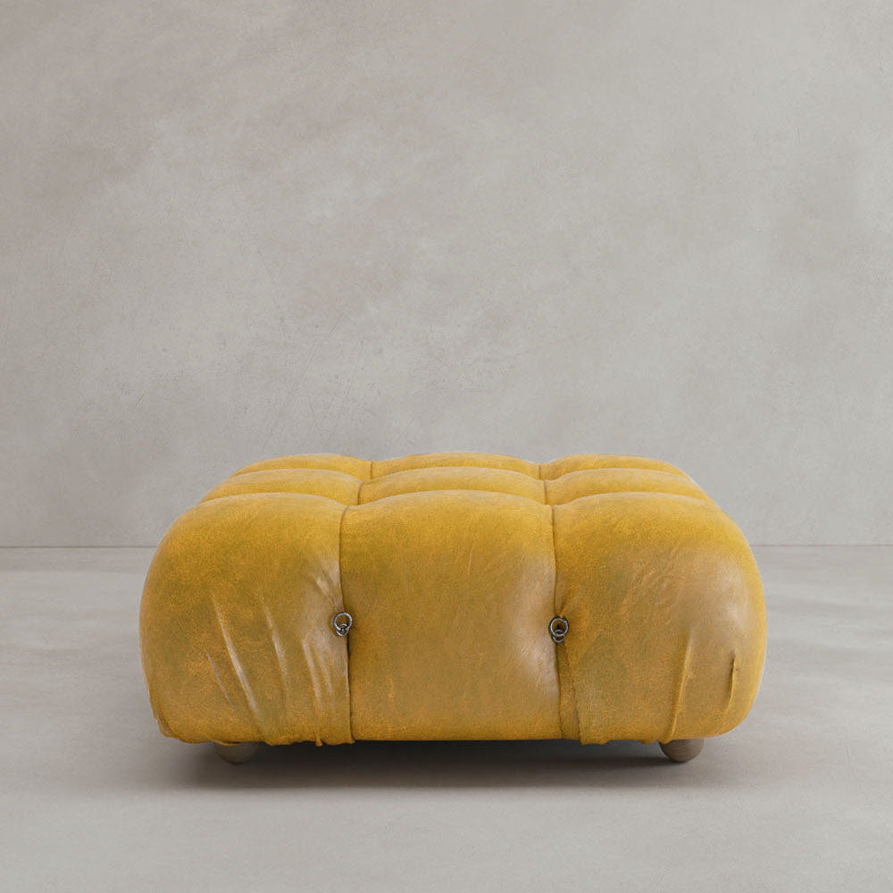 The Belmont Ottoman, a square tufted piece in mustard yellow with a cushioned surface, sits on a light gray floor against a plain wall, adding iconic classic charm to any modular design setting.