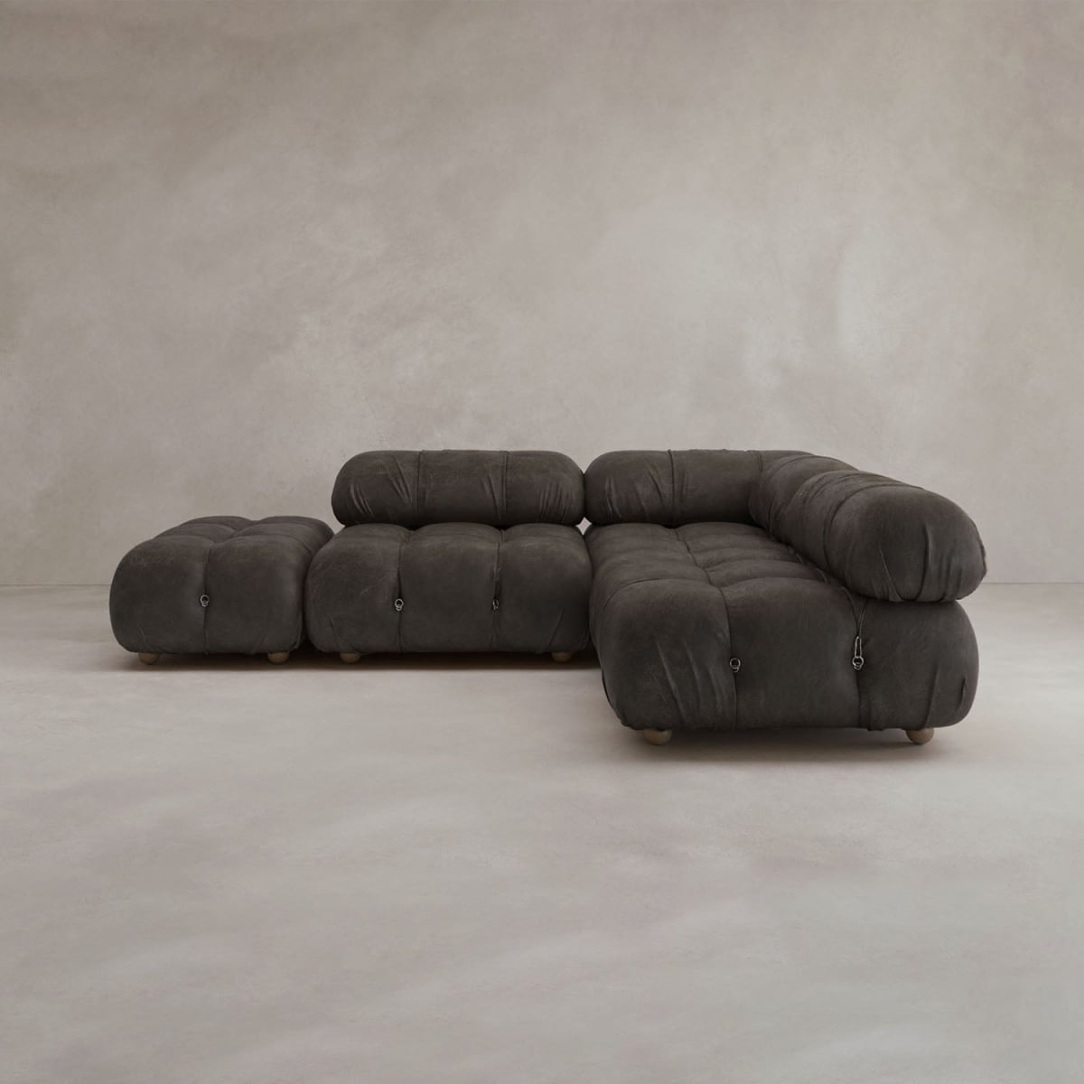 The Belmont Sofa Combination B is a modern, dark gray, tufted modular sofa with rounded cushions and low wooden legs, set in a minimalist room with light gray textured walls and floor.
