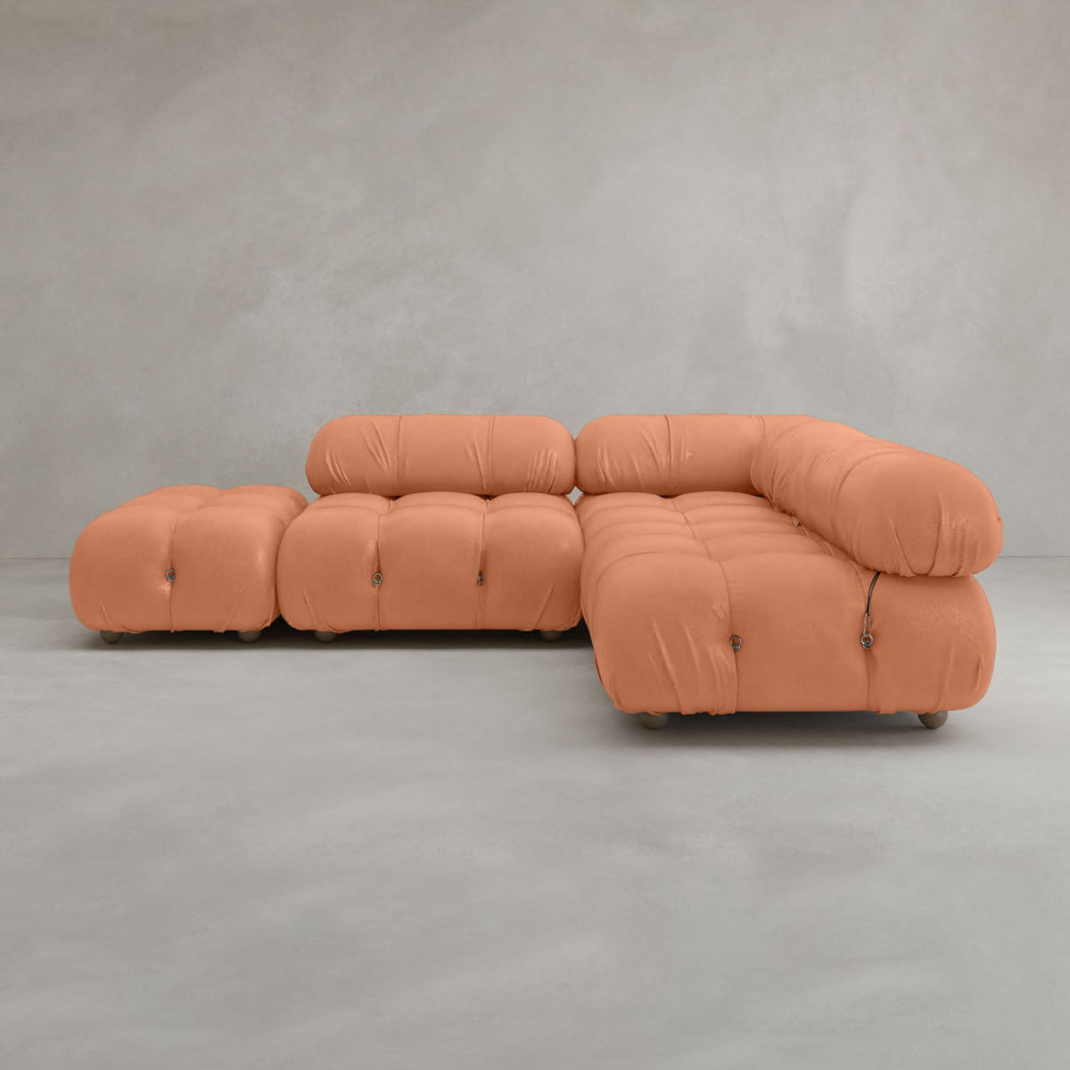 The Belmont Sofa Combination B is a modern, L-shaped modular sofa with tufted, puffy light orange upholstery. Set against a plain light gray wall and concrete floor, its low seat and armless design create a relaxed atmosphere.