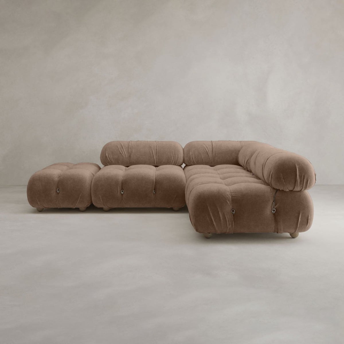 The Belmont Sofa Combination B features plush, rounded cushions in soft brown upholstery. This modern modular sofa includes a chaise lounge and ottoman, providing versatile comfort on a light gray floor against a textured neutral wall.