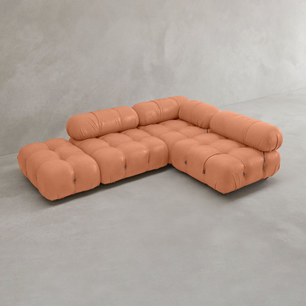 The Belmont Sofa Combination B, a modular tufted L-shaped orange sofa with low backrest and rounded cushions, sits on a light gray floor against a plain gray wall.