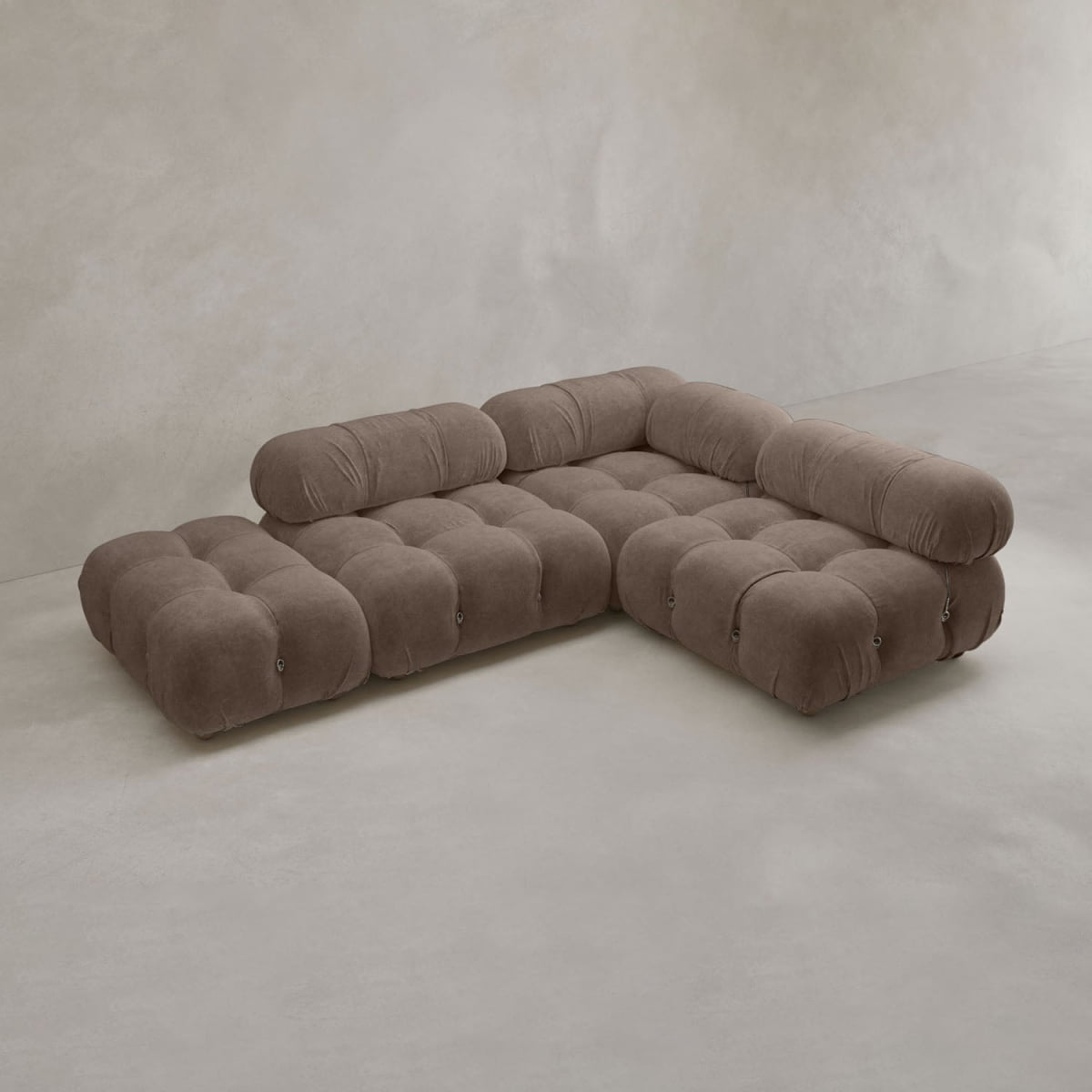 The Belmont Sofa Combination B is a modern, low-profile modular sofa featuring tufted, rounded cushions and brown upholstery, placed against a plain, light-colored wall and floor.
