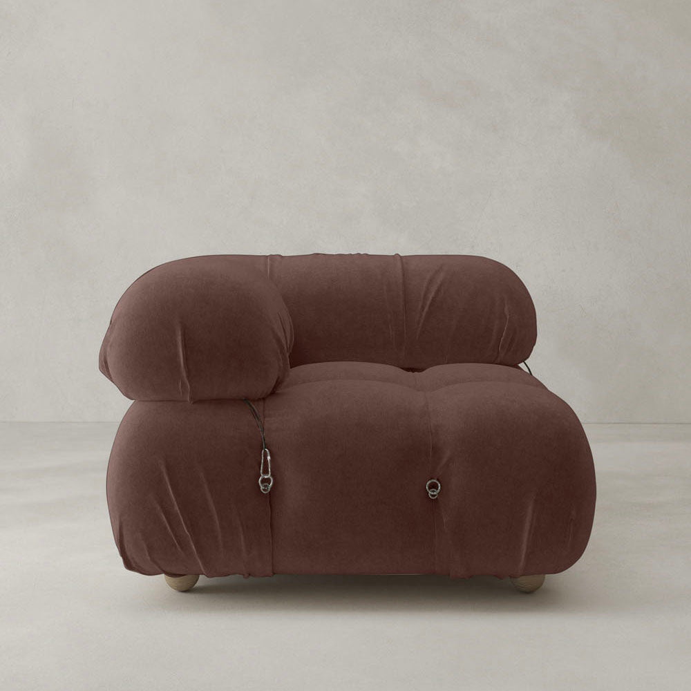 The Belmont Sofa Right Armrest is a modern, plush brown seat with rounded edges, soft upholstery, modular design, and visible metal buckles. It sits on a light gray floor against a plain off-white wall.