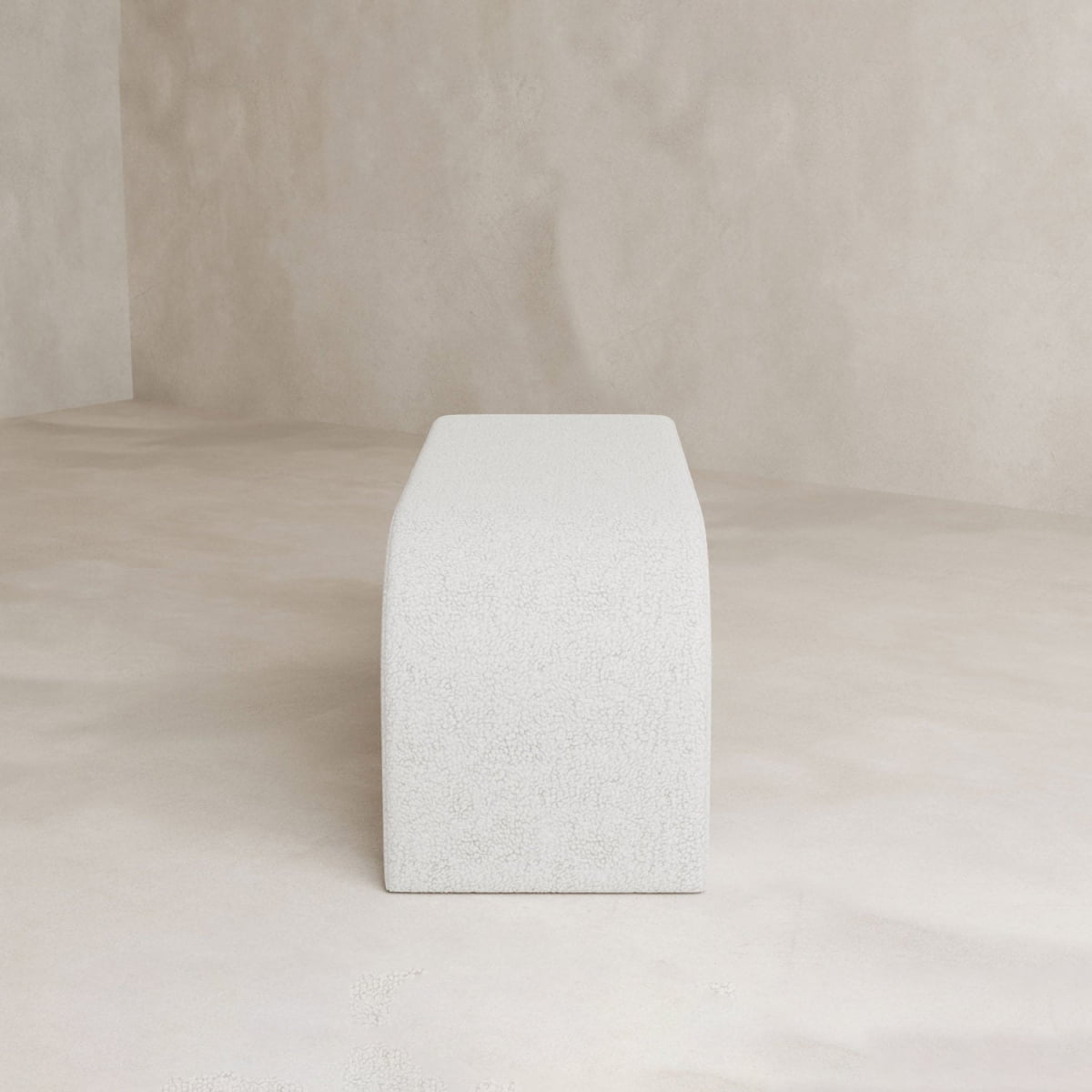The Beverly Bench, a minimalist rectangular white boucle piece with smooth, curved edges, sits in a neutral-toned room with textured walls and floor, adding modern elegance and refined style to the interior.