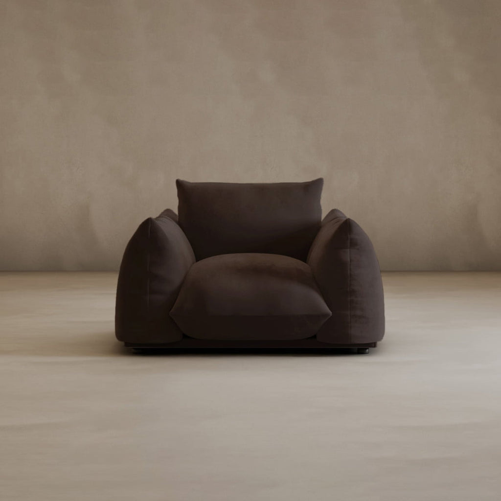 The large Calypso Arm Chair in dark brown features wide, plush cushions and sits on a light beige floor with a plain beige wall backdrop, creating an inviting and cozy look.