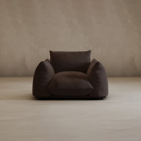 The large Calypso Arm Chair in dark brown features wide, plush cushions and sits on a light beige floor with a plain beige wall backdrop, creating an inviting and cozy look.