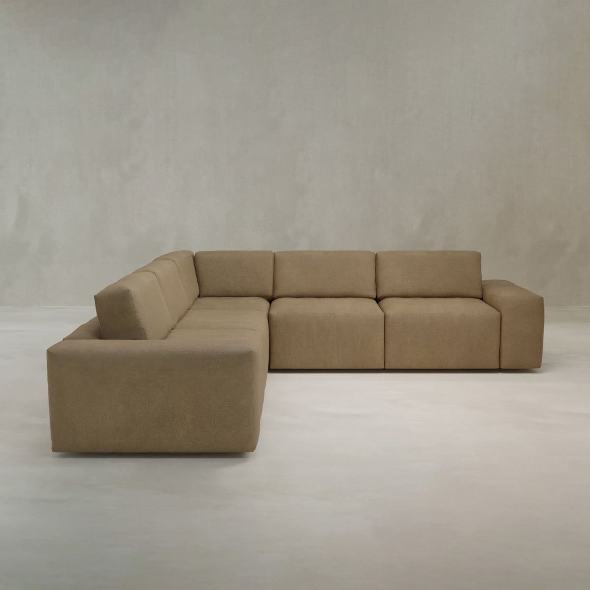 The Canonbury L Shape Sofa, a large modern sectional with wide blocky cushions and light brown high-performance fabric, sits in a minimalist room with neutral walls and flooring.