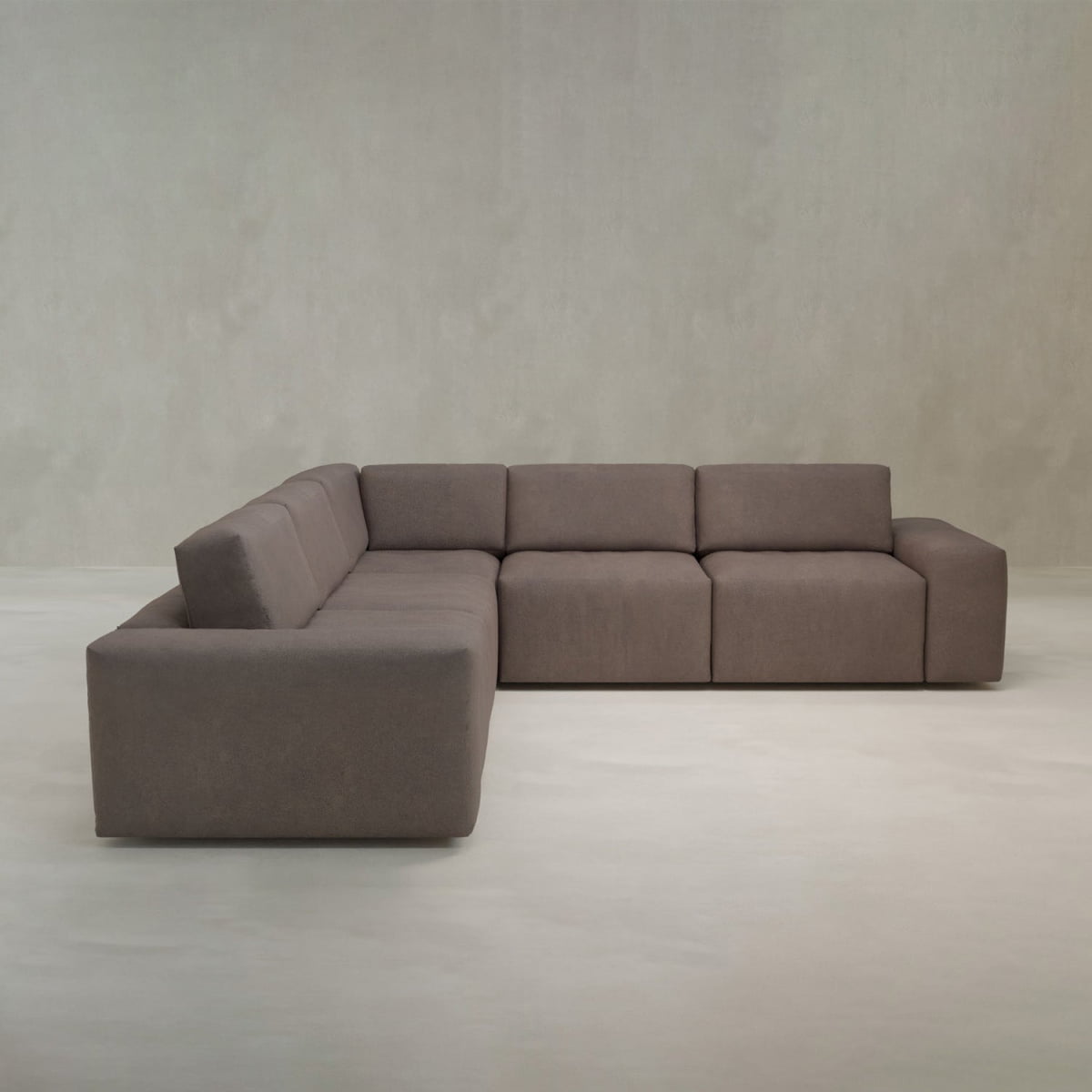The Canonbury L Shape Sofa features a modern, minimalist design with clean lines and boxy cushions, upholstered in high-performance brown fabric, set against a plain light grey wall and floor.