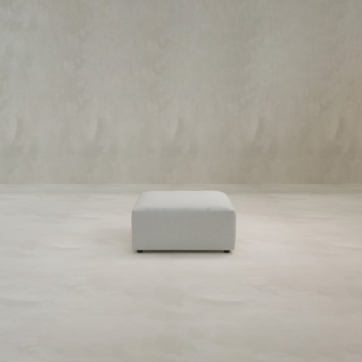 A minimalist scene featuring the Canonbury Ottoman in light gray, centered on a matching smooth floor with a textured neutral wall in the background.
