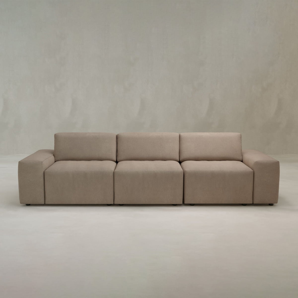 The Canonbury Three Seater Sofa features a modern, minimalist beige design with three wide seats and low backrests, delivering contemporary comfort in any space with light-colored walls and flooring.