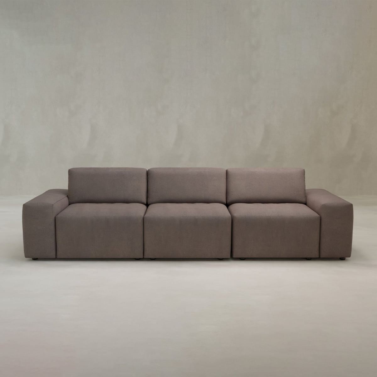 The Canonbury Three Seater Sofa features a modern, minimalist design with wide armrests, a low back, and high-performance plain taupe fabric upholstery, shown against a neutral light gray background for contemporary comfort.