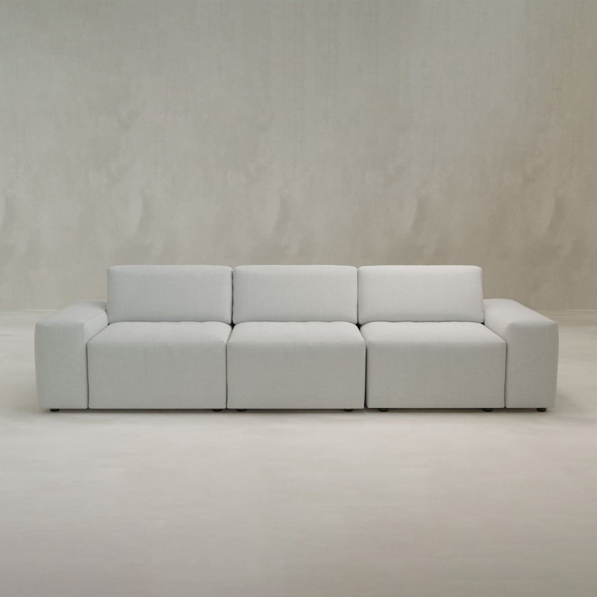 The Canonbury Three Seater Sofa, in light gray with wide seats and low backrests, enhances a minimalist room featuring matching pale gray walls and floors.