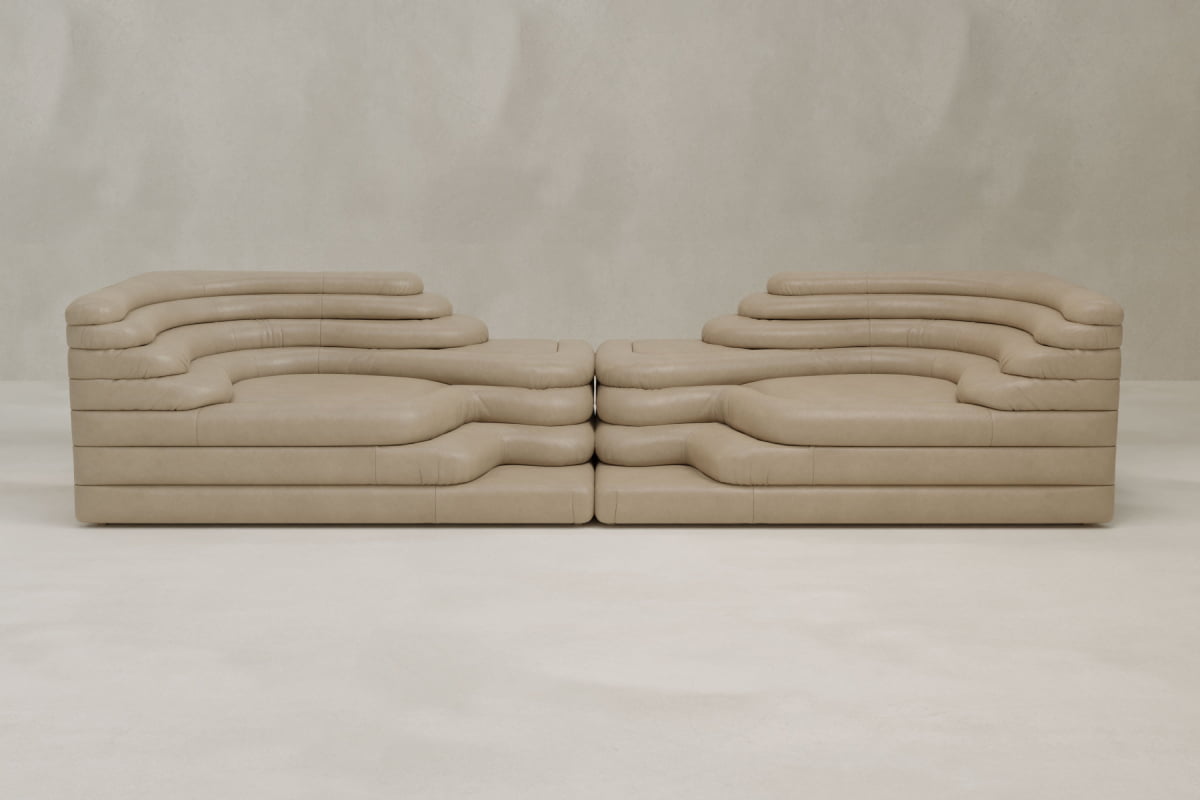 Two Cascata 2 Piece Sofas in beige leather, featuring modular design and wavy layered cushions, face each other, highlighting modern luxury against a light textured wall and floor.