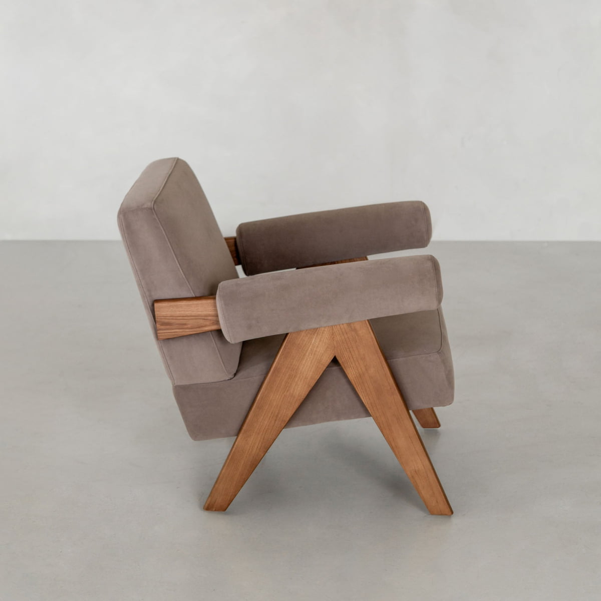 The Chelsea Lounge Chair features light brown faux mohair upholstery with angled wooden legs and armrests, embodying a mid-century style. It is positioned on a plain light gray floor against a minimalist, neutral background.
