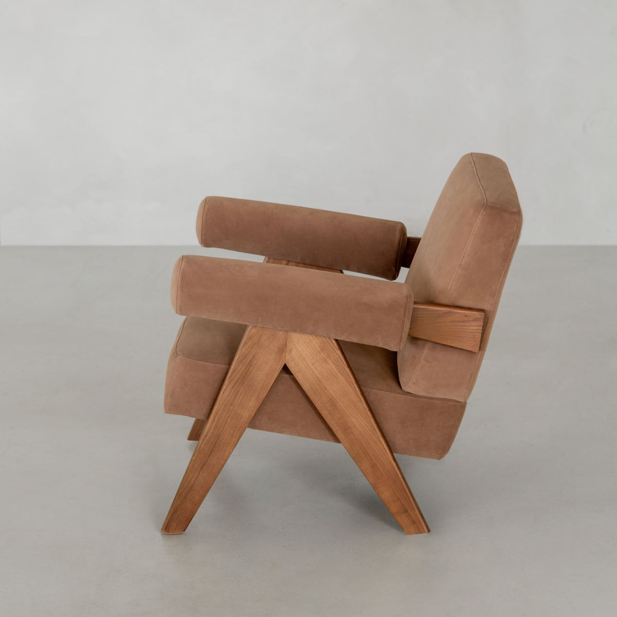 The Chelsea Lounge Chair has angled wooden legs, armrests, brown faux mohair cushions, and a minimalist mid-century look, shown on a light gray floor with a plain light wall behind.