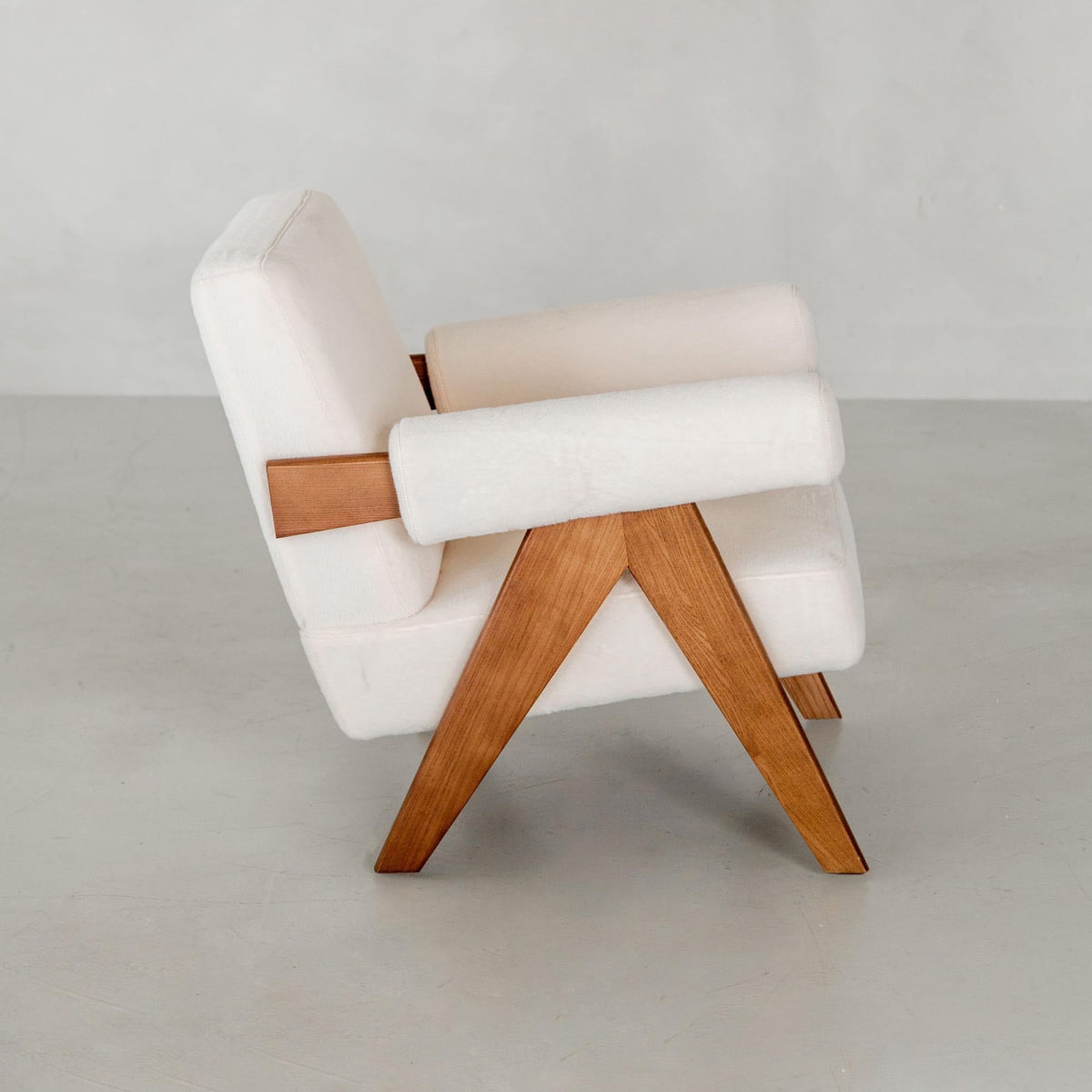The Chelsea Lounge Chair features white faux mohair upholstery with angled wooden legs and armrests, offering a refined mid-century look on a light gray floor against a plain background.