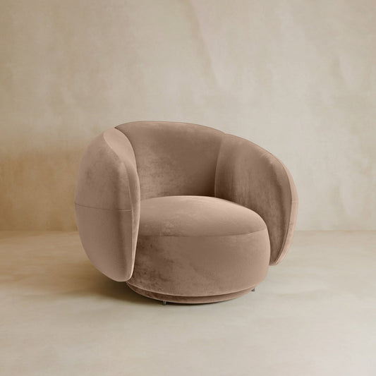 The Clara Armchair features beige velvet upholstery with a rounded back and sides, set against a neutral, textured background for contemporary style.