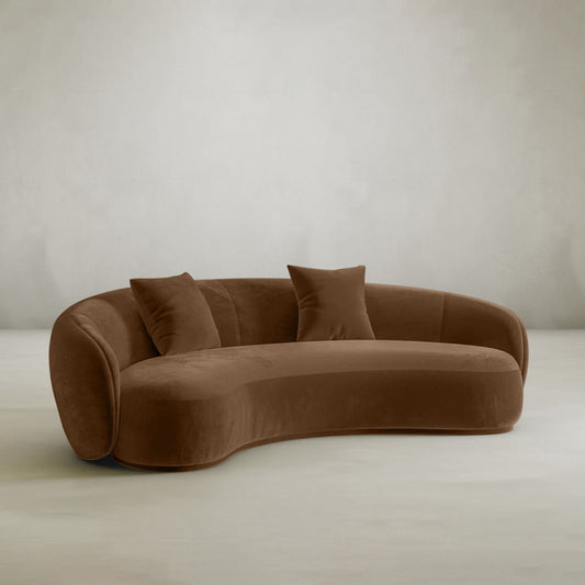 The Clara Sofa features a modern curved design with a smooth velvet finish and includes two matching pillows, displayed against a plain light background.