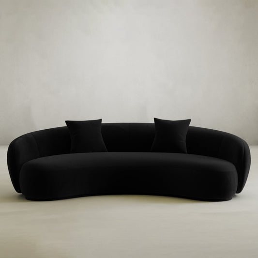 The Clara Sofa, a modern curved black piece with two matching throw pillows, adds style and comfort to any contemporary home against a neutral backdrop.