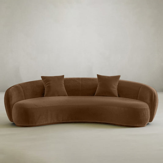 The Clara Sofa is a modern curved brown sofa with a velvety finish and two matching square pillows, bringing style and comfort to any contemporary home.
