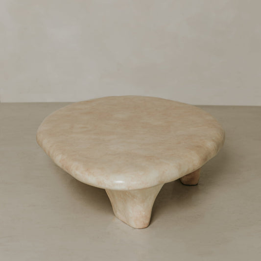 The Cleo Coffee Table is a round, low beige piece with three short, chunky legs, set against a neutral backdrop. It features subtle natural forms and hints of brutalist design for an avant-garde look.
