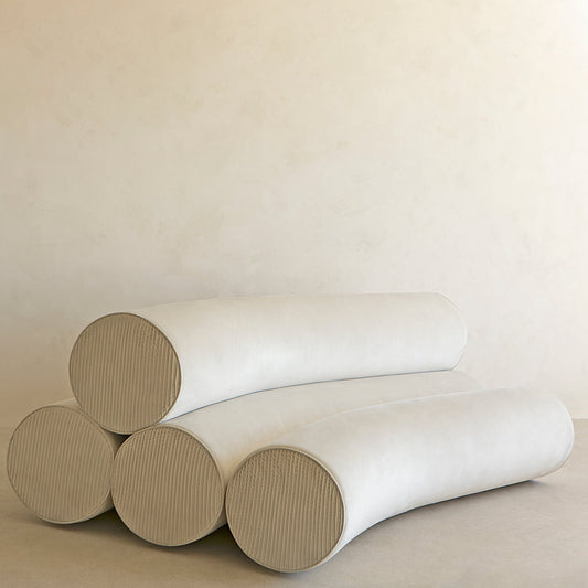 Four large, cylindrical, light beige cushions from the Cylindra Sofa Rental are stacked in a minimalist arrangement against an off-white wall, creating a sculptural and modern environment.