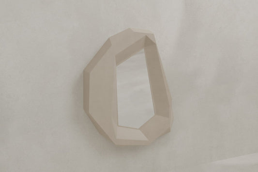The Facet Mirror Large features a geometric, irregular shape and a faceted beige frame, evoking the look of a cut gemstone. Its modern, brutalist design adds contemporary sophistication to any light-colored, textured wall.