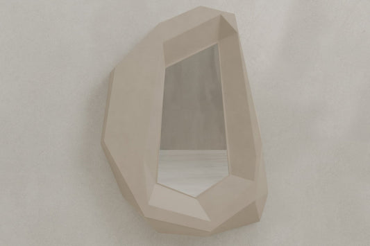 The Facet Mirror Large features a geometric, angular beige frame with brutalist style, resembling a faceted gemstone, and is displayed on a textured light gray wall.