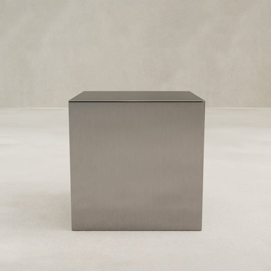 The Forge Steel Cube Plinth, featuring a brushed finish, showcases modern design and industrial flair as it rests on a smooth light floor against a neutral beige wall.