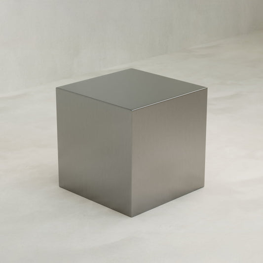 The Forge Steel Cube Plinth features smooth, reflective metallic surfaces and rests on a light gray, subtly textured floor with a matching backdrop, creating a modern look with understated industrial style.