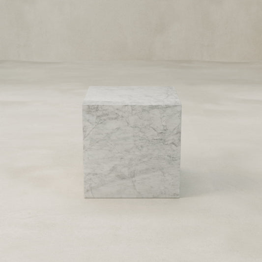 The Griselle Marble Plinth - Cube features a smooth, polished surface and rests on a neutral-toned floor—ideal for minimalist interiors with its solid marble construction and seamless look against a plain backdrop.
