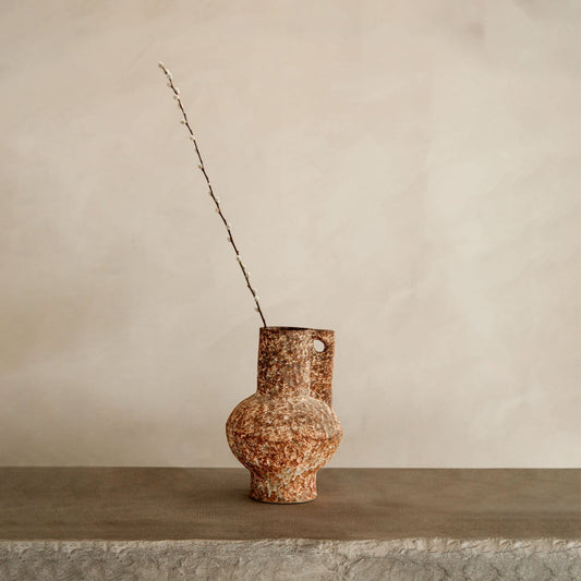 The Haru artisan pottery vase features a rustic, textured finish with a rounded base and narrow neck. Displayed with a thin branch on stone against a beige backdrop, its ideal for minimalist home decor.