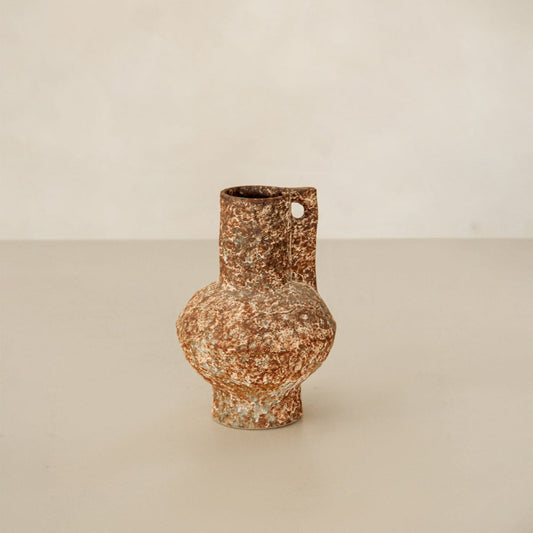 The Haru vase features a rustic, weathered metal finish with a rounded body, narrow neck, and small circular handle. Set on a neutral surface with a light beige background, it adds artisan pottery charm to your home decor.