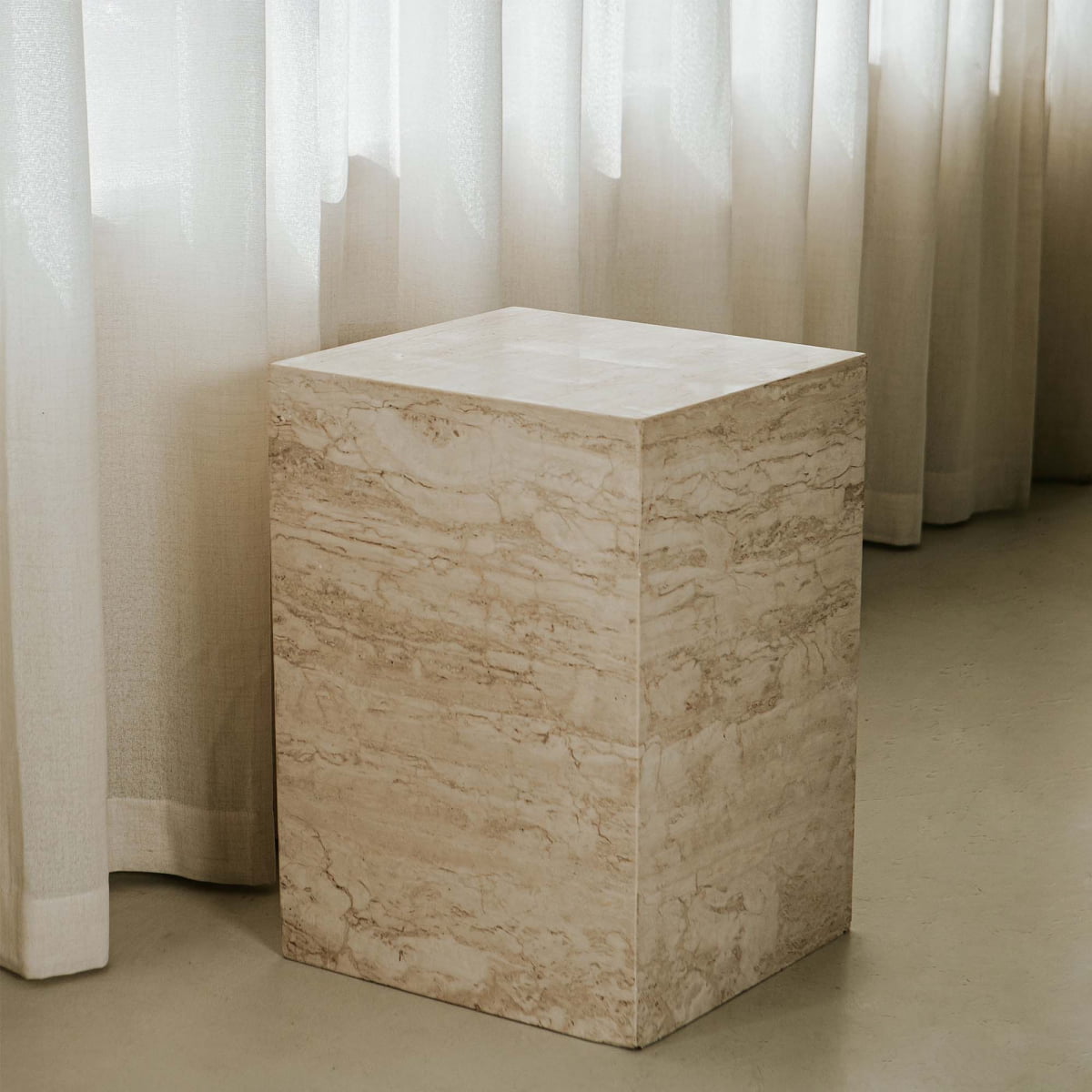 The Haven Travertine Plinth, featuring light beige and cream veining, sits on a smooth floor before sheer white curtains, adding an elegant decor touch to the space.