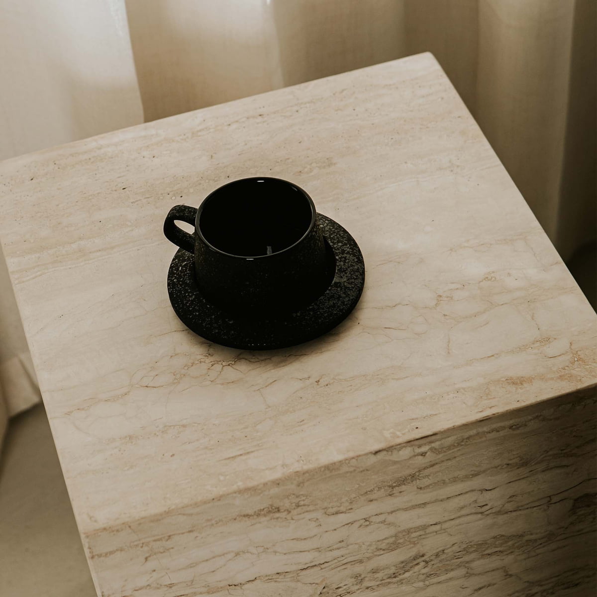 A black coffee cup and saucer rest on the Haven Travertine Plinth side table, adding elegant decor against soft, neutral curtains.