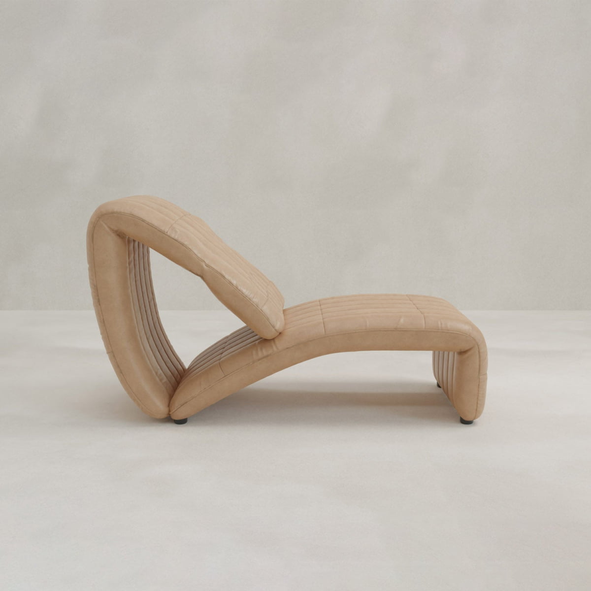 The Hillier Chair features a sculptural, curved design in premium vintage leather, offering smooth lines and a minimalist look. Its subtle reclining adds comfort, complemented by a neutral-toned floor and matching background.