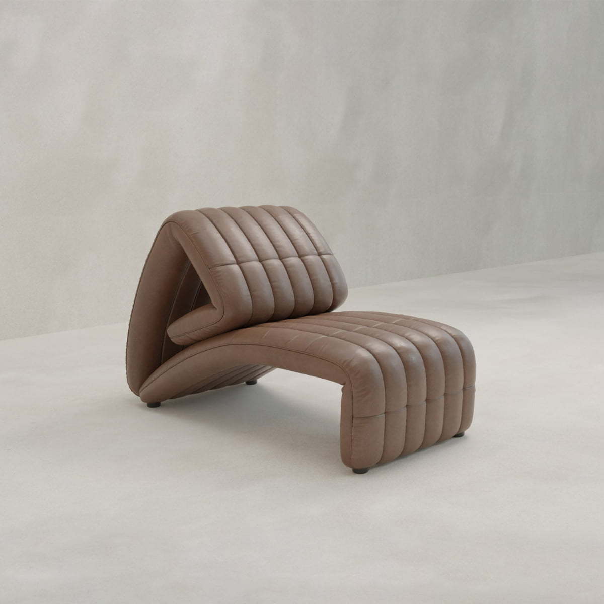 A Hillier Chair in premium vintage leather, with a curved wave design and vertical tufted lines, reclines for added comfort. It rests on a light floor beside a simple textured wall—an elegant touch for any modern lounge space.